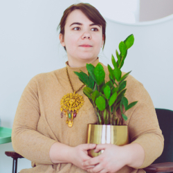 A photo of a white woman with dark hair, looking off into the distance while holding a ZZ plant in a gold pot.