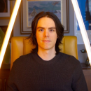 A photo of a white man with medium long dark hair. He is sitting in a yellow chair and is lit up with dramatic blue and yellow light tubes. 